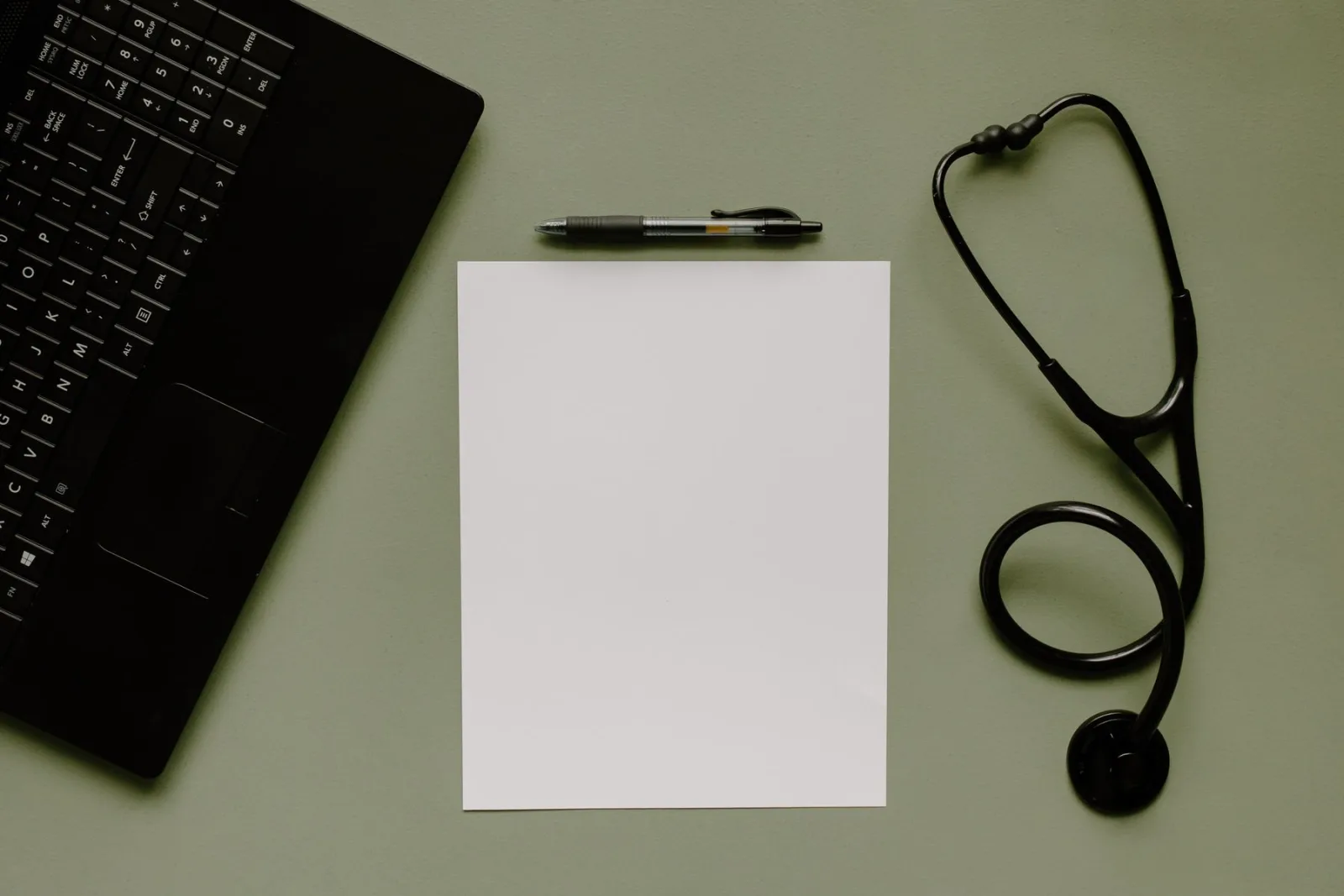 A blank sheet of paper, pen and stethoscope beside a laptop.